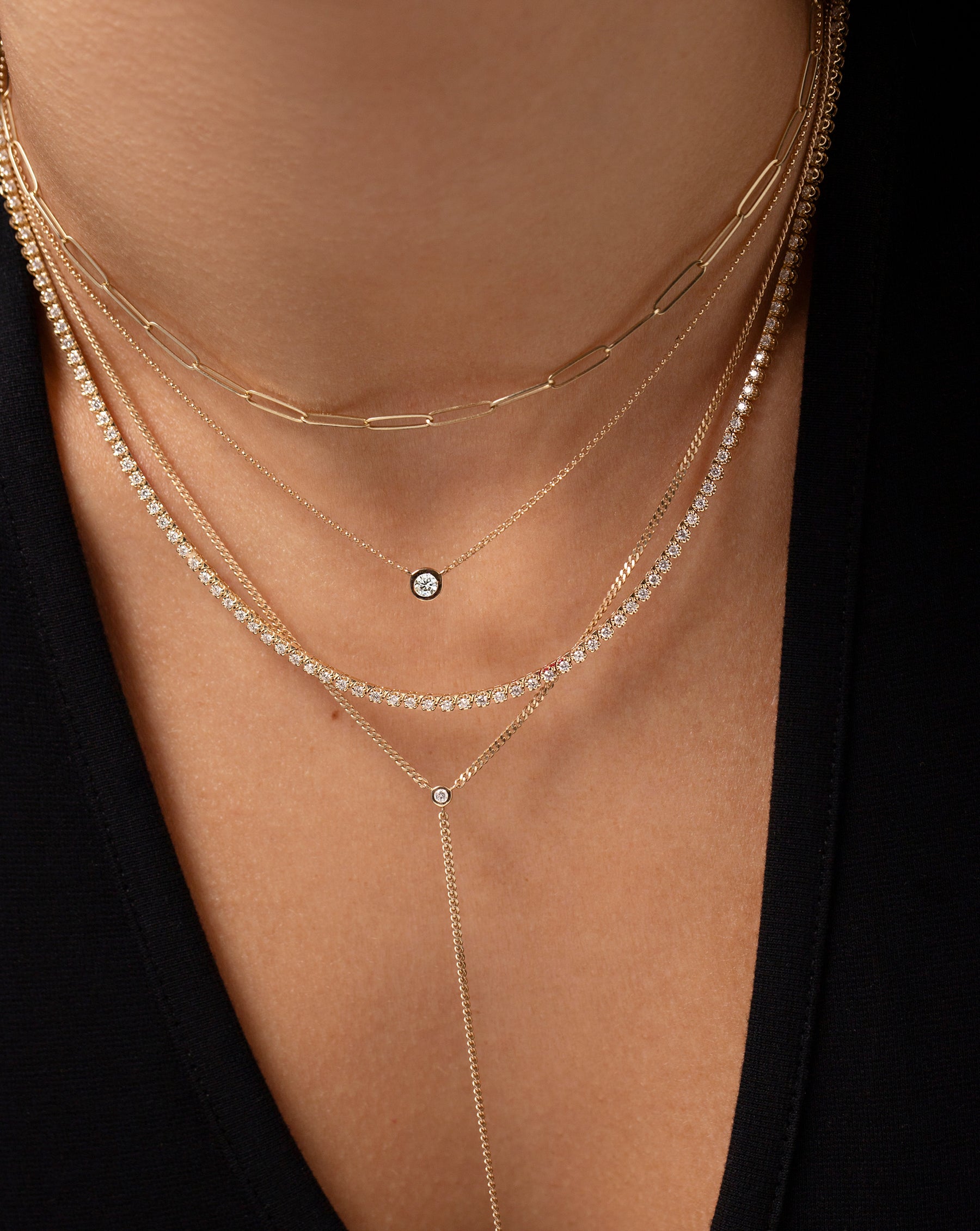 close up image of 14k yellow gold link chain necklace, bezel-set round diamond pendant necklace, classic diamond tennis necklace, and lariat chain necklace shown on model